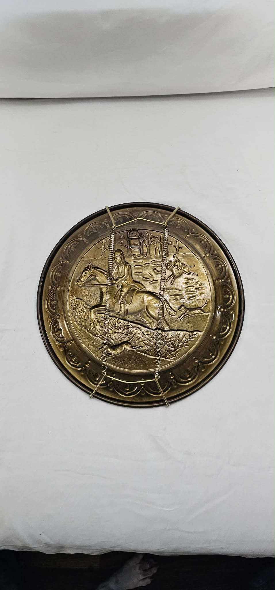 Single vintage brass-tone hunting-scene wall plate with a mounted rider and hounds, shown with the spring-style hanger attached.