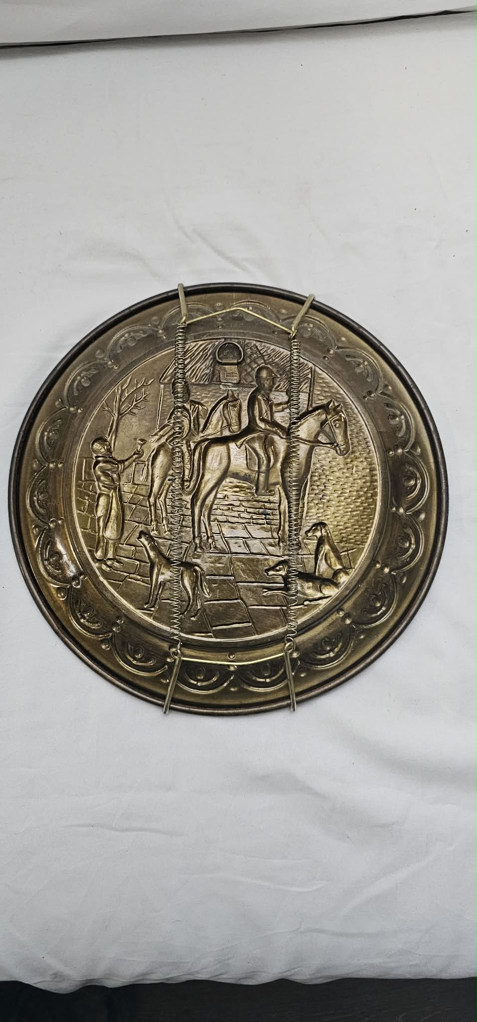Back view of a vintage embossed hunting-scene wall plate showing the spring-style hanger and aged brass-tone finish.
